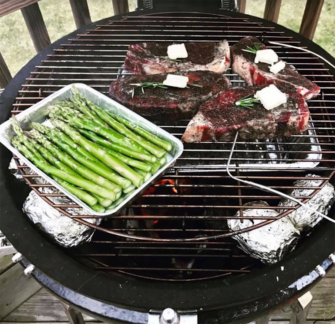 steaks on a grill with potatoes wrapped in foil and asparagus in an aluminum tray