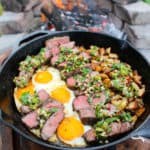 a skillet next to a fire filled with fried eggs, sliced steak, and potatoes topped with charred scallion chimichurri