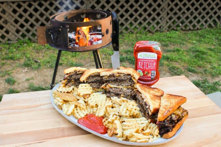 large platter on a table in front of a grill filled with cheesy patty melt sandwiches, fries, and ketchup