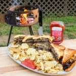 large platter on a table in front of a grill filled with cheesy patty melt sandwiches, fries, and ketchup