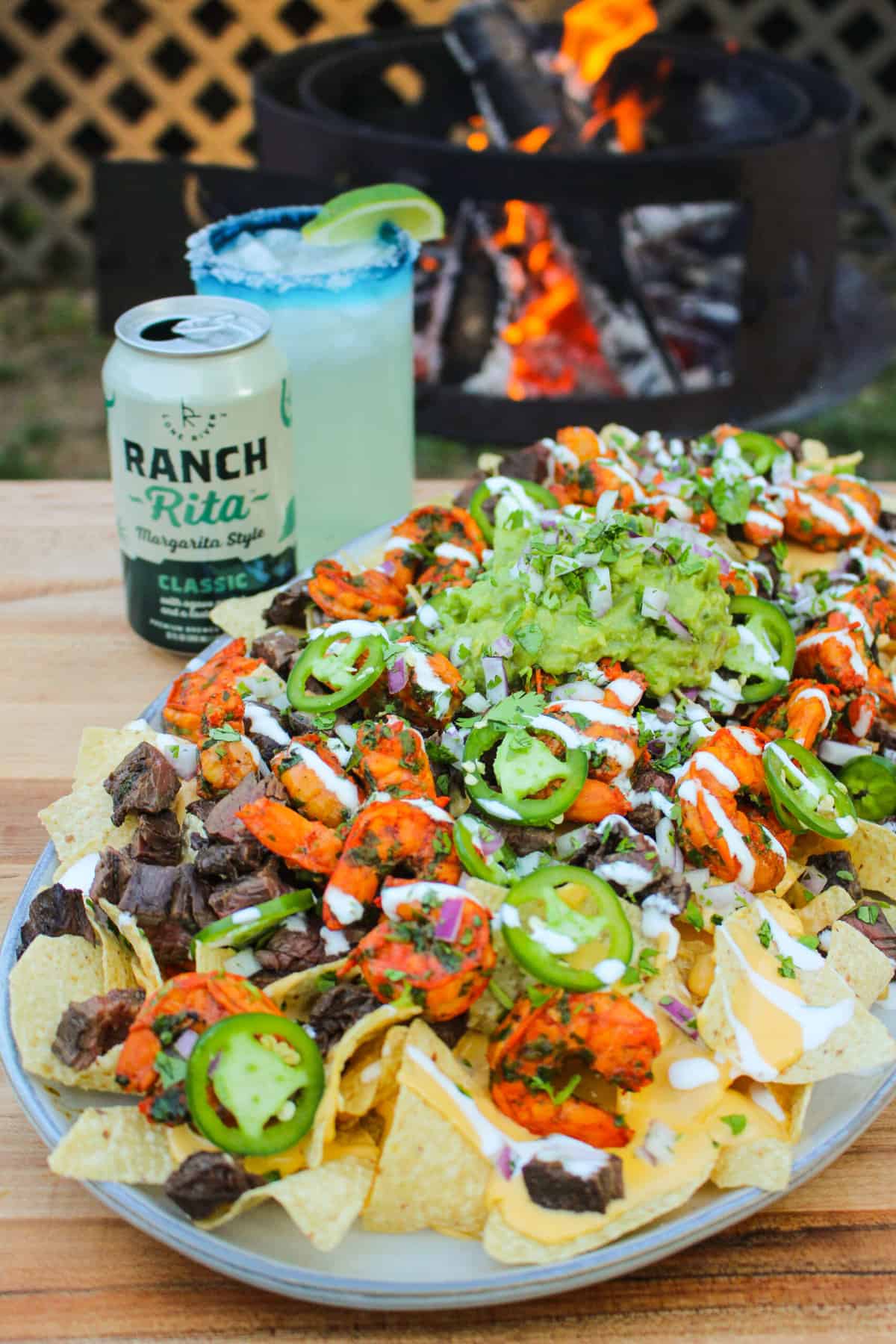 closeup of a platter full of nachos with a margarita and grill in the background