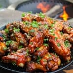 a large plate piled with huli huli chicken wings and garnished with sesame seeds, sriracha, and cilantro