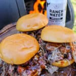 side view of three pulled pork sandwiches next to a smoker and a can of Ranch Water