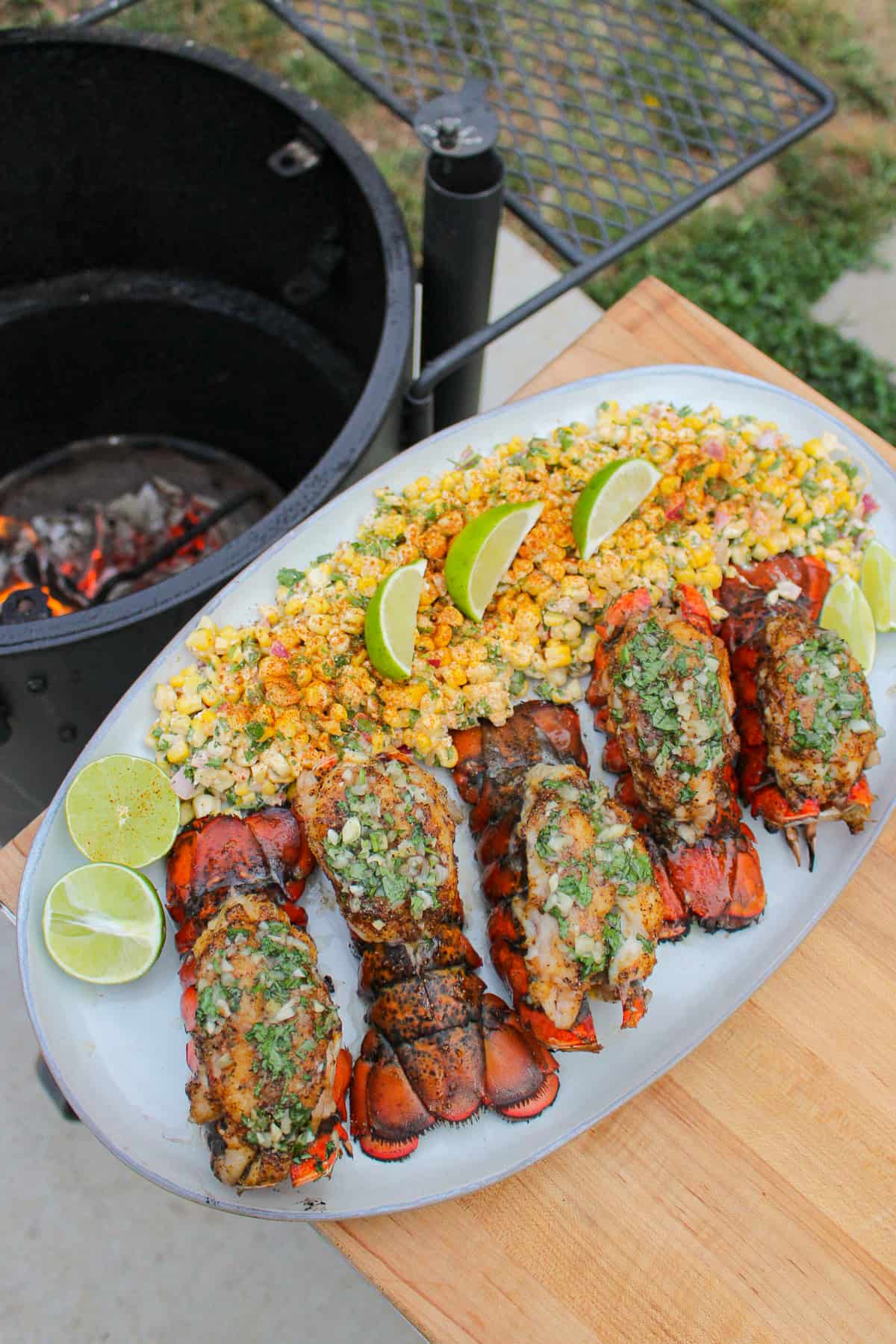 finished smoked lobster tails with esquites on a white plate next to a smoker